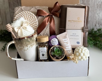 May include: A gift basket filled with self-care items. It includes a mug, herbal tea, a journal, chocolate, a candle, a succulent, and bath products. The items are arranged in a white box and tied with a brown ribbon.