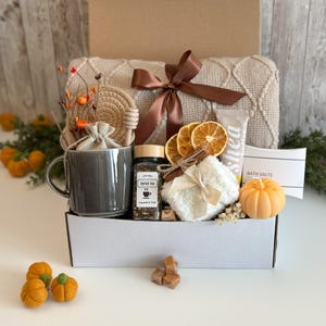 K&ouml;nnte beinhalten: Eine Geschenkbox mit einem grauen Becher, Kr&auml;utertee, einem kleinen Handtuch, Badesalz und einem dekorativen K&uuml;rbis. Die Box enth&auml;lt auch eine beige Decke, getrocknete Orangenscheiben und ein braunes Band. Das Farbschema ist warm und herbstlich.