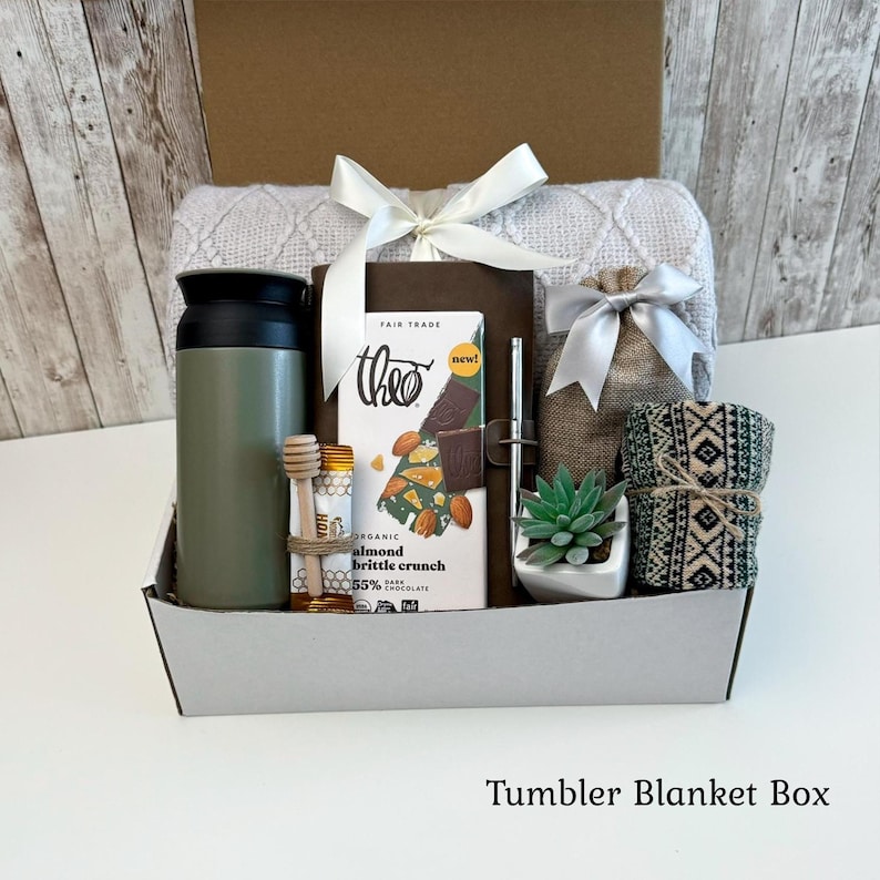 May include: A gift box containing a sage green tumbler, a Theo almond brittle crunch chocolate bar, a wooden honey dipper, a silver pen, a small succulent in a white pot, a burlap pouch, and a patterned fabric roll, all topped with a white ribbon.