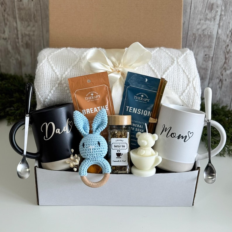 May include: A gift box containing a white textured blanket with a ribbon, a black mug labeled Dad, a white mug labeled Mom, two silver spoons, a blue crochet rabbit rattle, a jar of herbal tea, and a bear-shaped candle in a small cup.
