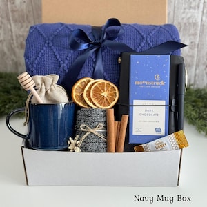 May include: A gift box containing a blue knitted blanket with a navy ribbon, a blue ceramic mug with a wooden honey dipper, a small beige pouch, dried orange slices, grey socks, cinnamon sticks, a black notebook with a pen, and Moonstruck dark chocolate.