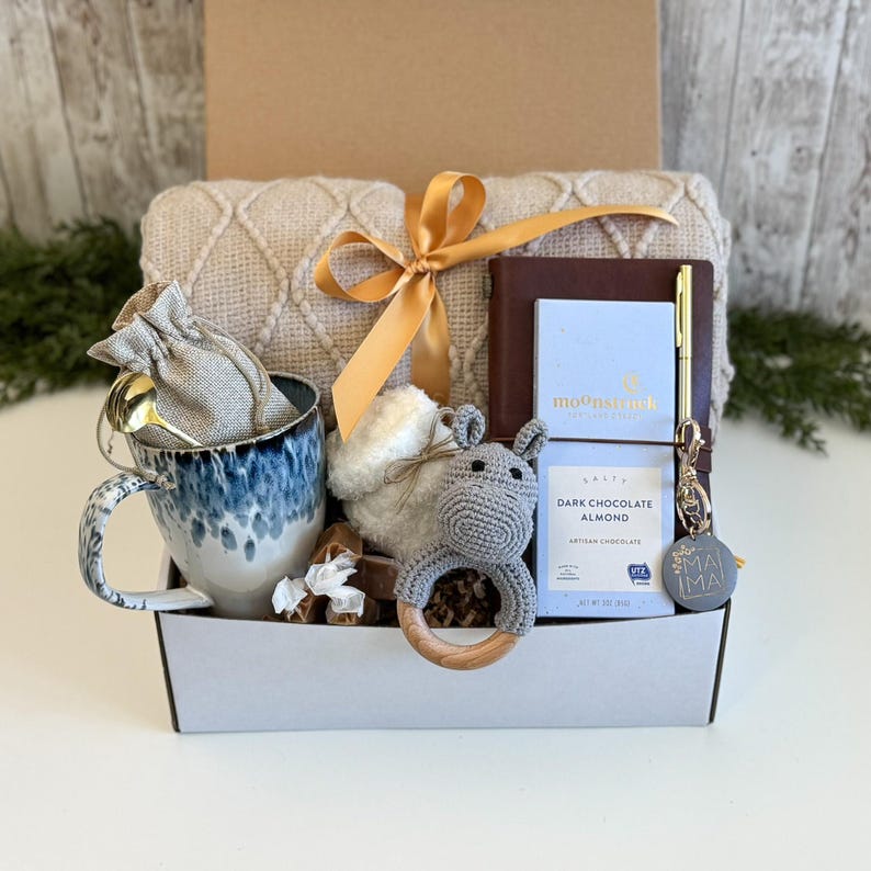 K&ouml;nnte beinhalten: Eine Geschenkbox mit einer cremefarbenen, strukturierten Decke mit goldener Schleife, einem blau-wei&szlig; gesprenkelten Keramikbecher, einem kleinen Leinenbeutel, einer grauen H&auml;kelrassel, einem dunkelbraunen Notizbuch, einem goldenen Stift und dunkler Schokolade.