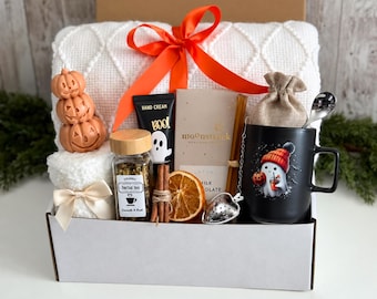 May include: A gift box containing a white textured blanket with an orange ribbon, a stack of three orange pumpkin soaps, herbal tea, hand cream, a chocolate bar, cinnamon sticks, a dried orange slice, a tea infuser, and a black mug with a ghost graphic.