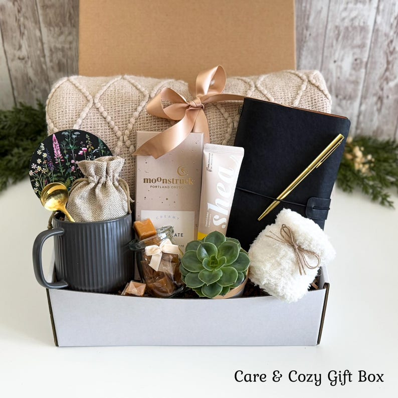 May include: A gift box containing a beige knit blanket, a black journal with a gold pen, a Moonstruck chocolate bar, a shea cream tube, a gray ribbed mug with a gold spoon, a small burlap pouch, a glass jar of caramels, and a green succulent.