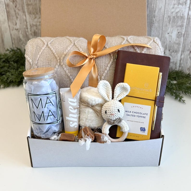 K&ouml;nnte beinhalten: Eine Geschenkbox mit einer beigen, strukturierten Decke mit goldener Schleife, einem Glas mit der Aufschrift MAMA, einer Tube Sheacreme, einer wei&szlig;en H&auml;kel-Hasenrassel mit Holzring, einem dunkelbraunen Notizbuch, einem Goldstift und Milchschokolade.