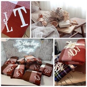 May include: A collection of personalized red and white plaid blankets with various embroidered initials. The blankets are folded and stacked on a table with a neon sign that reads "In the bag Design".