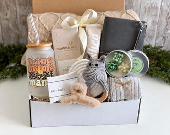 May include: A gift box containing a frosted glass tumbler with a leopard print mama graphic, a Moonstruck chocolate bar, a shea cream tube, a dark notebook, a succulent candle, a gray crochet hippo toy, a beige hair claw, and gray socks.