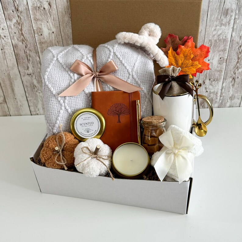 K&ouml;nnte beinhalten: Ein Geschenk-Set mit herbstlichen Artikeln. Es enth&auml;lt eine wei&szlig;e Decke, ein braunes Lederjournal, eine Duftkerze, eine Tasse mit goldenem Griff und dekorative K&uuml;rbisse. Das Set wird in einer wei&szlig;en Box pr&auml;sentiert.