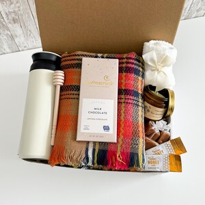 K&ouml;nnte beinhalten: Eine Geschenkbox gef&uuml;llt mit einem cremefarbenen isolierten Becher, einem h&ouml;lzernen Honigl&ouml;ffel, einem karierten Schal, einer Tafel Milchschokolade, einer kleinen Kerze, verpackten Karamellen und Honigp&auml;ckchen. Die Box ist braun.