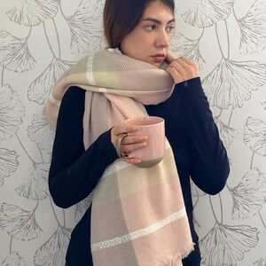 May include: A soft, oversized scarf featuring a large plaid pattern in shades of blush pink, cream, and muted beige, accented with thin white horizontal stripes and finished with a delicate fringed edge, paired with a solid pink ceramic mug.