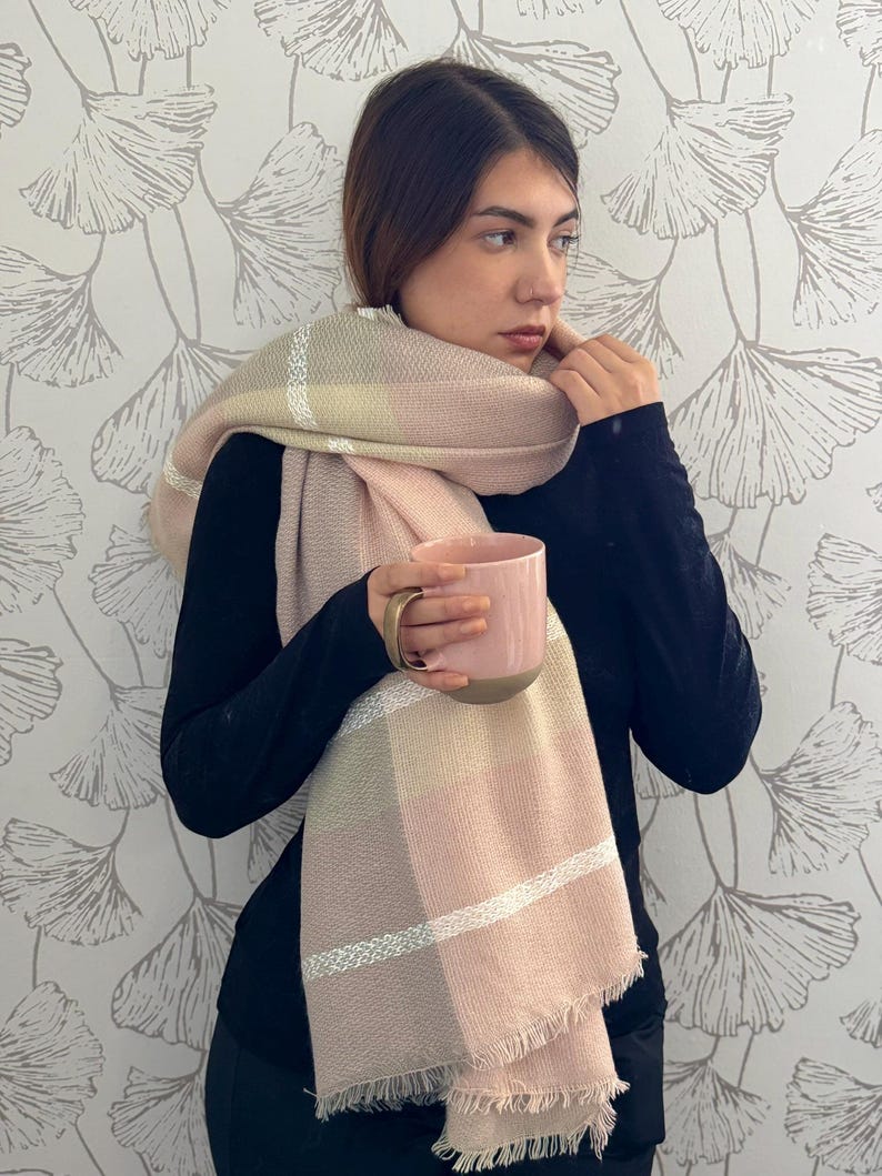 May include: A soft, oversized scarf featuring a large plaid pattern in shades of blush pink, cream, and muted beige, accented with thin white horizontal stripes and finished with a delicate fringed edge, paired with a solid pink ceramic mug.