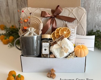 May include: A gift box containing a grey ceramic mug, a jar of herbal tea, a white fuzzy item, a tube of shea butter, bath salts, a small orange pumpkin, dried orange slices, and a beige knit blanket tied with a brown ribbon.