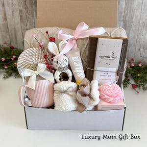 May include: A gift box containing a pink ribbed mug with a white bow, a silver spoon, a crochet rabbit rattle, a tube of shea cream, a brown notebook with a Moonstruck milk chocolate bar, a pink flower-shaped candle, and a white fuzzy item.