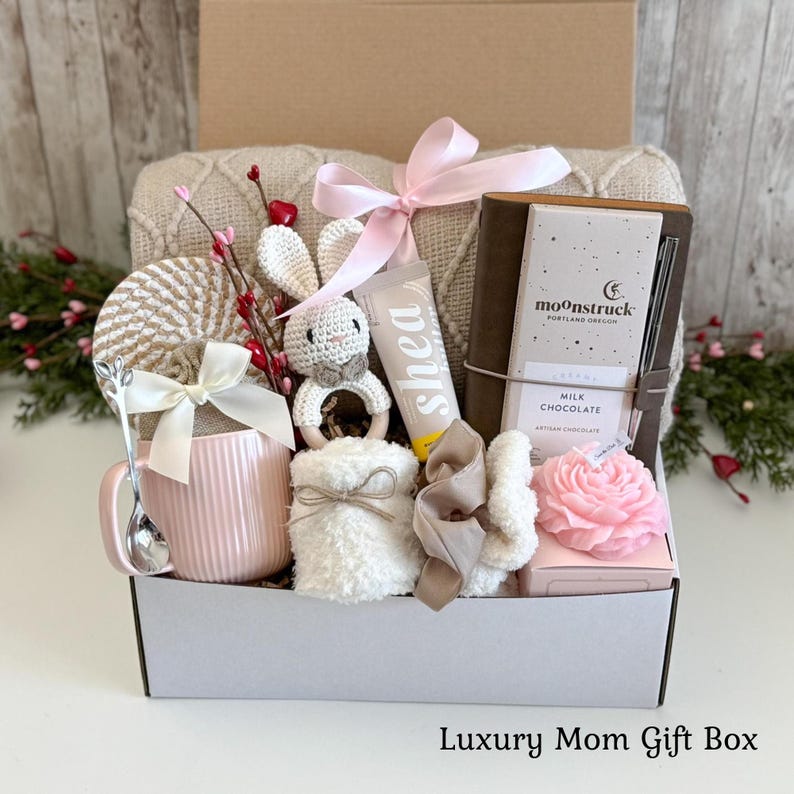 May include: A gift box containing a pink ribbed mug with a white bow, a silver spoon, a crochet rabbit rattle, a tube of shea cream, a brown notebook with a Moonstruck milk chocolate bar, a pink flower-shaped candle, and a white fuzzy item.