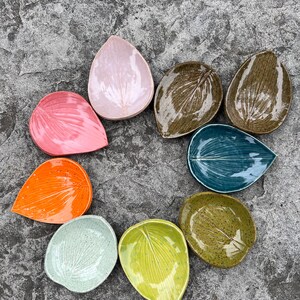May include: A collection of nine ceramic dishes shaped like leaves, arranged in a circular pattern. The dishes feature various glossy finishes in colours including light pink, dark brown, teal, olive green, lime green, mint, orange, and red.