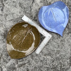 May include: Two leaf-shaped ceramic dishes, one olive green and one periwinkle blue, both featuring speckled glaze and embossed vein textures. A metal ruler is placed between them, showing the olive dish is approximately 10 cm in length.