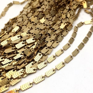 Puede incluir: Primer plano de una cadena de oro hecha de pequeños eslabones rectangulares. Los eslabones tienen una superficie texturizada y estampada.