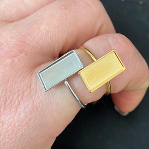 May include: Two silver and gold rings with rectangular settings. The rings are on a finger.