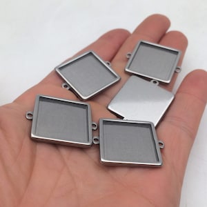 May include: Five silver-toned square metal pendant trays with a smooth, shiny finish. Each tray has two small loops on the top for attaching a chain or cord.