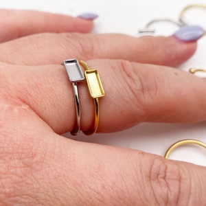 Może przedstawiać: Dwa pierścionki z prostokątnymi oprawami. Pierścionek po lewej stronie jest srebrny, a pierścionek po prawej stronie jest złoty.