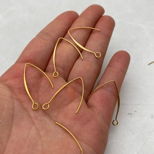 Può includere: Gancetti per orecchini dorati, noti anche come fili per orecchini, sono mostrati su una mano. I gancetti sono in metallo e hanno un design semplice e curvo.