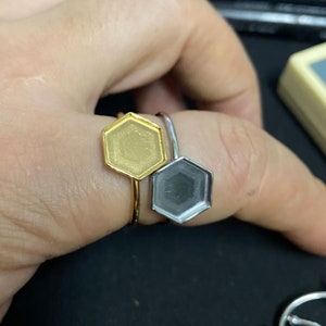 May include: Two hexagon-shaped rings, one gold and one silver, are shown on a finger. The rings are simple and have a smooth, flat surface.