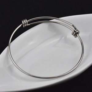 May include: A silver-coloured, expandable bangle bracelet. The bracelet is made of a thin, round metal and features a sliding mechanism for adjustment. The bracelet is resting on a white, oval-shaped dish against a black background.