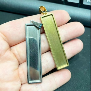 Może przedstawiać: Dwa prostokątne tace na wisiorki, jedna posrebrzana i jedna złota, trzymane w dłoni. Wisiorki mają prostokątną ramkę z pętlą na górze na łańcuszek. Złoty wisiorek ma złotą pętlę.