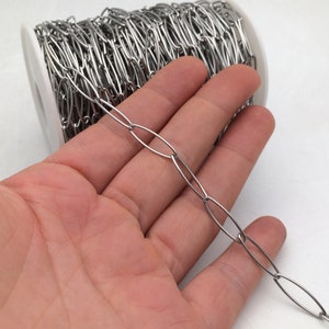 以下が含まれることがあります： 銀色の楕円形のチェーンが手に載せられています。チェーンは背景のスプールに巻き付けられています。