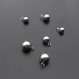 Puede incluir: Una colección de pequeños colgantes esféricos de metal plateado brillante con bucles adjuntos. Los colgantes están dispersos sobre una superficie gris oscuro, reflejando la luz. Estos colgantes son adecuados para la fabricación de joyas o proyectos de artesanía.