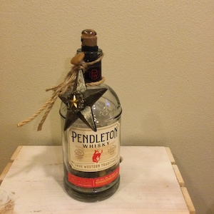 May include: A clear glass Pendleton Whisky bottle with a star-shaped decoration and rope accents. The bottle has a cork stopper and a label with the words "Pendleton Whisky" and a red bull logo. The bottle is sitting on a white wooden surface.