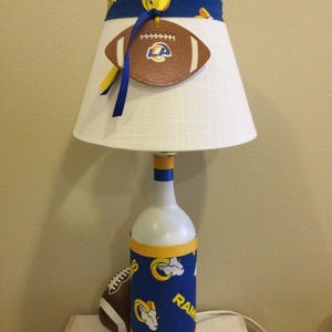 May include: A white lamp with a lampshade featuring a football-themed design. The lamp base is a repurposed bottle, adorned with blue and yellow fabric. A decorative football hangs from the lampshade. A football is placed next to the lamp.