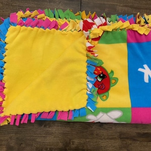 May include: A colorful fleece tie blanket with a yellow square in the center and a cartoon strawberry character on the right side. The blanket has a knotted fringe border in pink, blue, green, and yellow.