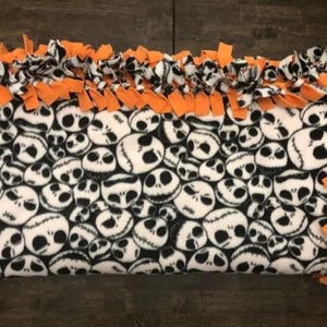 May include: A fleece tie blanket with a black and white pattern of Jack Skellington's face from the Nightmare Before Christmas. The blanket has a knotted fringe border in orange.