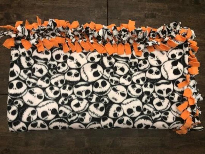 May include: A fleece tie blanket with a black and white pattern of Jack Skellington's face from the Nightmare Before Christmas. The blanket has a knotted fringe border in orange.
