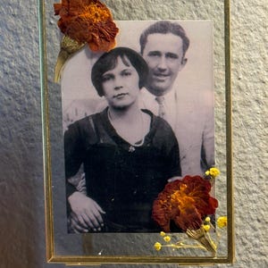 May include: A black and white photo of a couple in a gold frame with dried flowers. The woman is wearing a dark dress and the man is wearing a suit. The flowers are red and yellow.