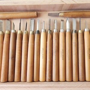 May include: A collection of wood carving tools with light-colored wooden handles and metal blades, arranged in a wooden box. Several tools are marked with the text Skillman Japan. The set includes various blade shapes for detailed woodworking.