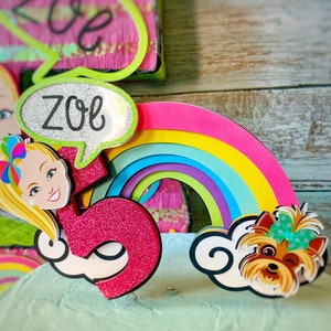 May include: A pink and green cake topper with a rainbow, a number 5, a cartoon girl with blonde hair, and a cartoon dog with a pink bow. The cake topper says "Zoe" in a speech bubble.