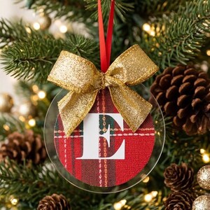 Puede incluir: Un adorno navideño circular de acrílico transparente con un inserto de tela de cuadros rojos y verdes y una letra E mayúscula blanca impresa en el centro. El adorno está rematado con un lazo de cinta dorada brillante y una cinta roja.