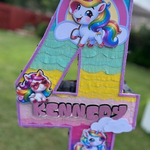May include: A colorful number 4 piñata decorated with unicorn graphics and the name "KENNEDY". The piñata features a rainbow color scheme, with a large unicorn at the top and smaller unicorns on the front.