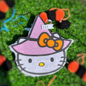 Puede incluir: Una piñata de Hello Kitty rosa y blanca con un sombrero de bruja y un lazo naranja. El sombrero tiene brillantina y el nombre "ANA" está escrito en una etiqueta blanca.