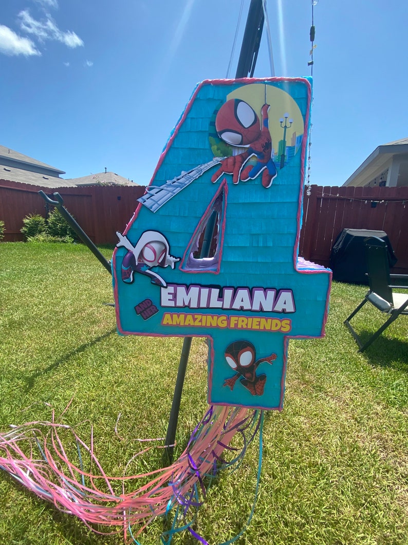 Może przedstawiać: Niebieska i r&oacute;żowa pi&ntilde;ata w kształcie cyfry 4, ozdobiona postaciami Spidermana. Pi&ntilde;ata ma napis "EMILIANA AMAZING FRIENDS".