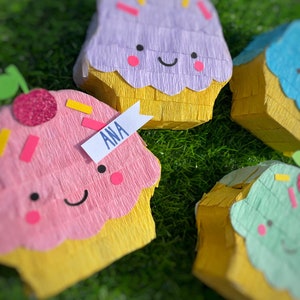 Puede incluir: Un conjunto de piñatas con forma de cupcake en colores pastel. Las piñatas tienen glaseado rosa, morado, azul y verde con bases amarillas. Cada una tiene una cara sonriente, chispas y una cereza. Una piñata tiene una etiqueta que dice "ANA".