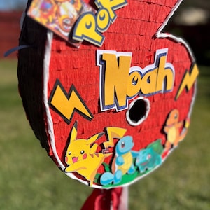 Puede incluir: Piñata roja con forma de número seis, decorada con personajes de Pokémon y el nombre "Noah" en letras amarillas. La piñata presenta a Pikachu, Squirtle y Charmander. La palabra "Pokémon" también es visible.