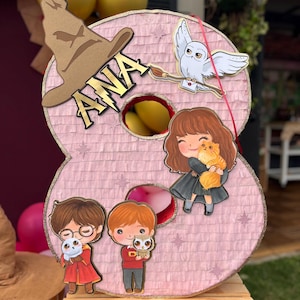 Puede incluir: Una piñata rosa con forma de número ocho decorada con un sombrero seleccionador marrón, un búho blanco y tres personajes de dibujos animados. El nombre ANA está impreso en letras negras sobre el sombrero. La piñata tiene estrellas y aberturas.