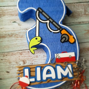 May include: A blue and brown number three piñata with a fishing theme. The piñata has a fishing rod, a fish, a tackle box, and the name "Liam" on it.