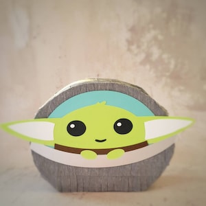 Puede incluir: Un personaje verde de Grogu, con grandes ojos negros y una pequeña sonrisa, está acunado en una cuna gris con forma de papel. La cuna tiene un interior azul claro y una banda marrón. El diseño recuerda a una piñata.