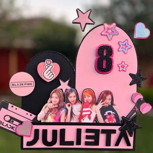 Puede incluir: Un adorno para tarta rosa y negro con el nombre "JULIETA" en letras grandes. El adorno presenta imágenes de cuatro personas, estrellas, corazones y el número 8. También se incluye una cinta de cassette y un micrófono.