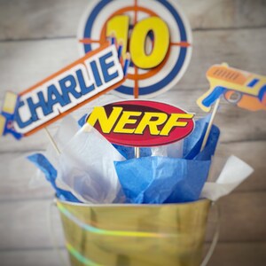 May include: A gold bucket filled with blue and white tissue paper. There are several party decorations on top of the tissue paper, including a red and yellow "NERF" sign, a blue and red target, a blue and white "CHARLIE" sign, and a toy gun.