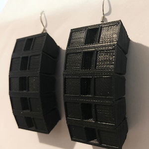 May include: A pair of black, geometric earrings. Each earring features stacked, rectangular blocks with cut-out details, suspended from silver-coloured ear wires. The earrings have a textured surface and a modern, architectural design.